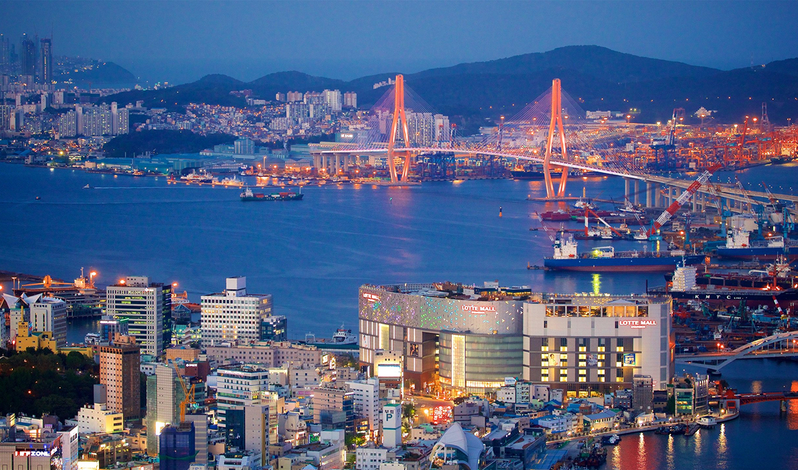 Vista da cidade de busan, bastante iluminada de cima, da pra ver o mar, um shopping, navios e várias casas e prédios.